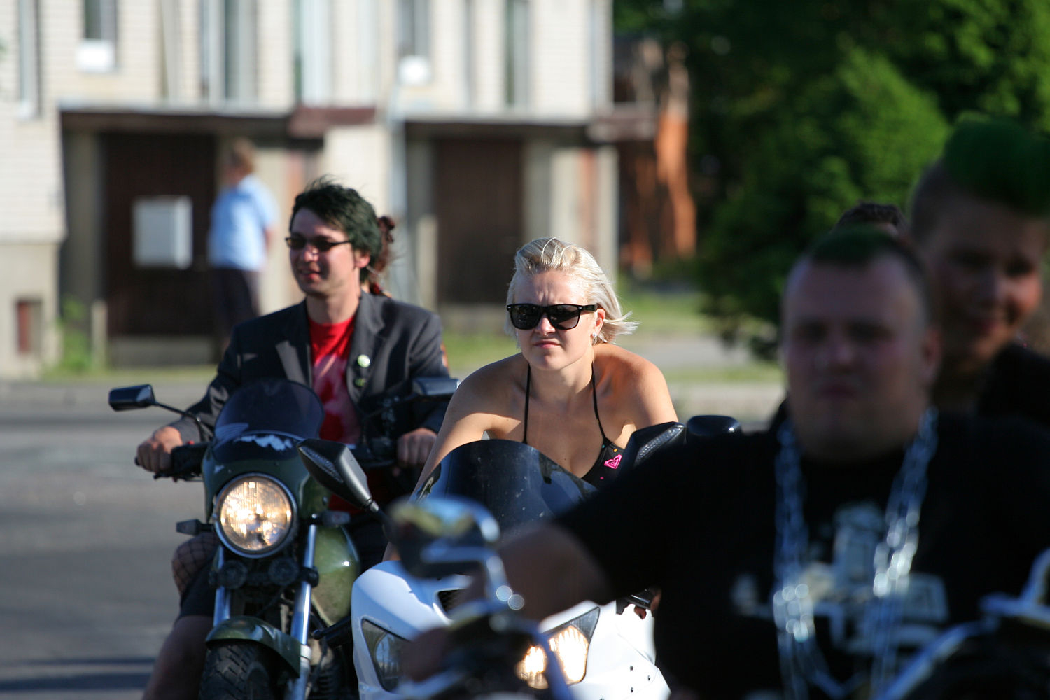 A group of motorcyclists rides down a street on a sunny day. At the front, a punk woman in sunglasses and a black sleeveless top rides a white motorcycle. A man next to her wears a dark jacket and sunglasses on a black motorcycle. Green trees and buildings are in the background, contrasting with their edgy style.