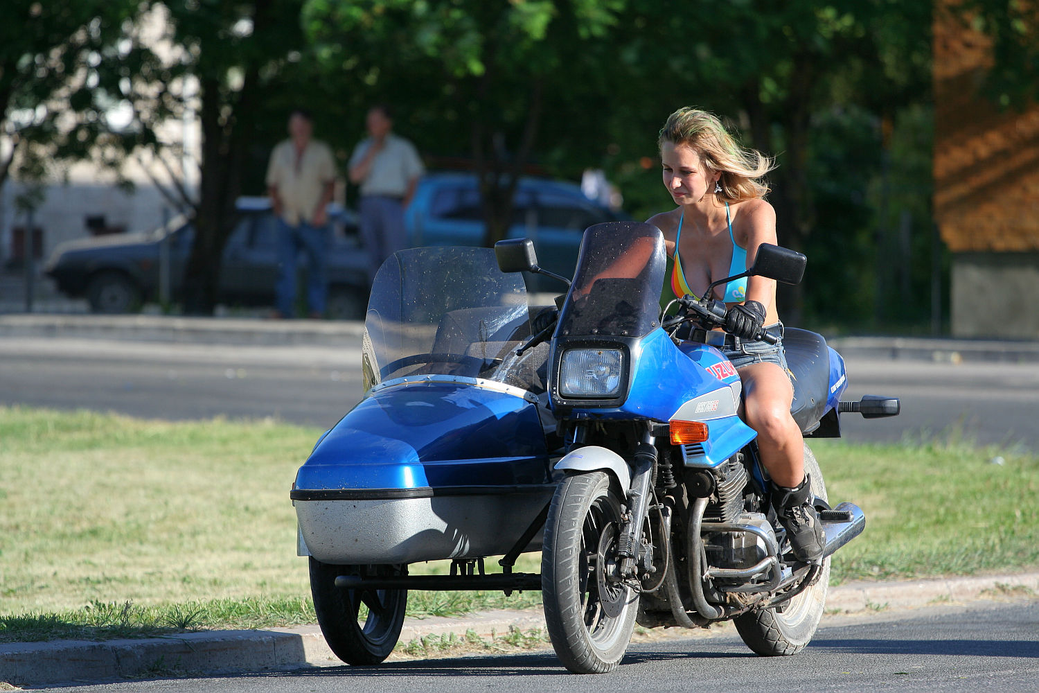 A woman with blonde hair, wearing a punk-inspired blue top and black shorts, rides a punk-style blue motorcycle with a sidecar on a paved road. The background is blurred with trees and two pedestrians, suggesting motion. The scene feels energetic as if capturing the spirit of Laulupidu during daylight.