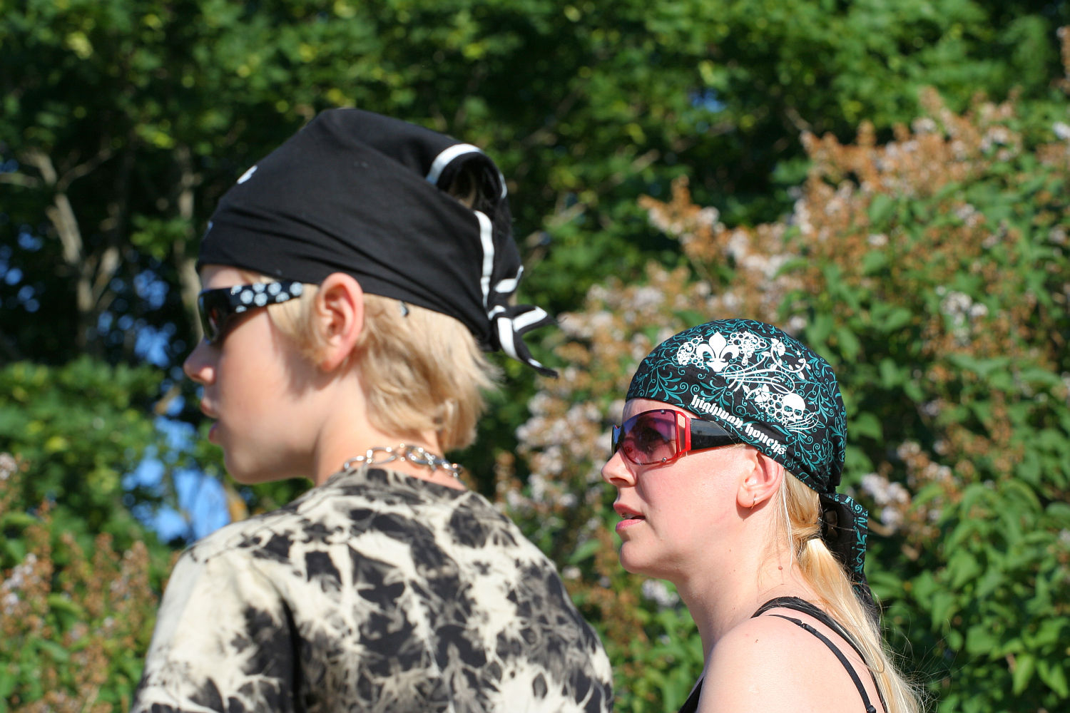 Two people outdoors on a sunny day, both wearing black bandanas and sunglasses. One sports a black and white tie-dye shirt with a chunky necklace, while the other rocks a punk-inspired black tank top. Lush green foliage surrounds them, reminiscent of Laulupidu's natural beauty. They seem focused on something off-camera.