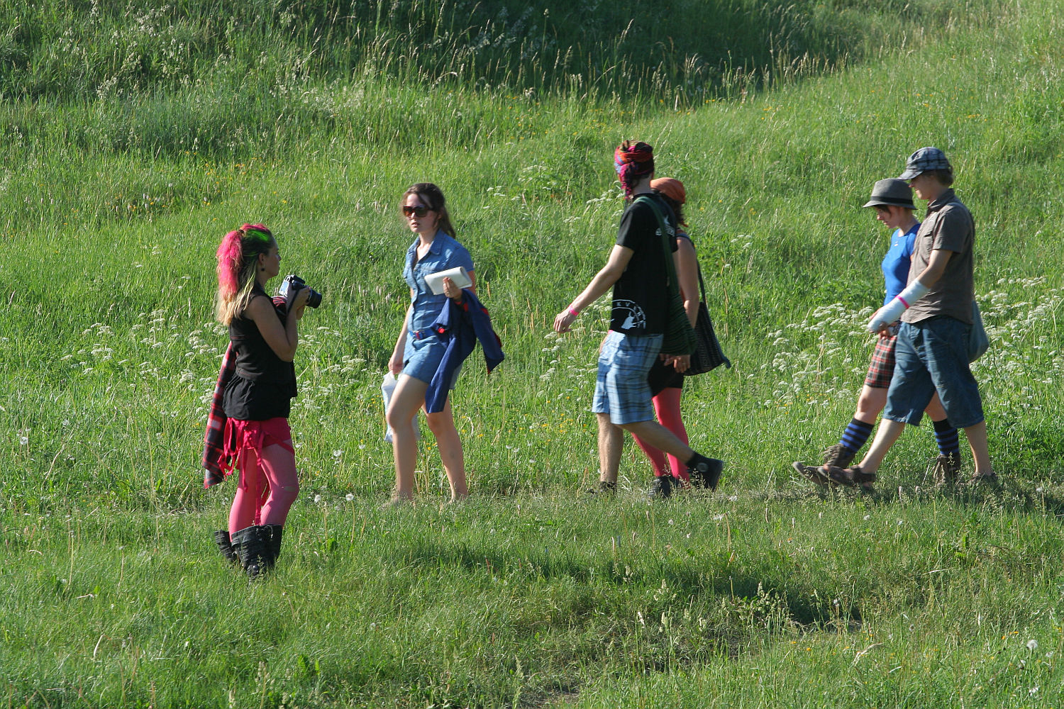 A group of people, five adults, are walking through a grassy field on a sunny day. They are dressed in a mix of casual and colorful attire, with some wearing hats and bright leggings reminiscent of Punk Laulupidu festivity. One person is holding a camera and appears to be taking photos. The field is lush and green.