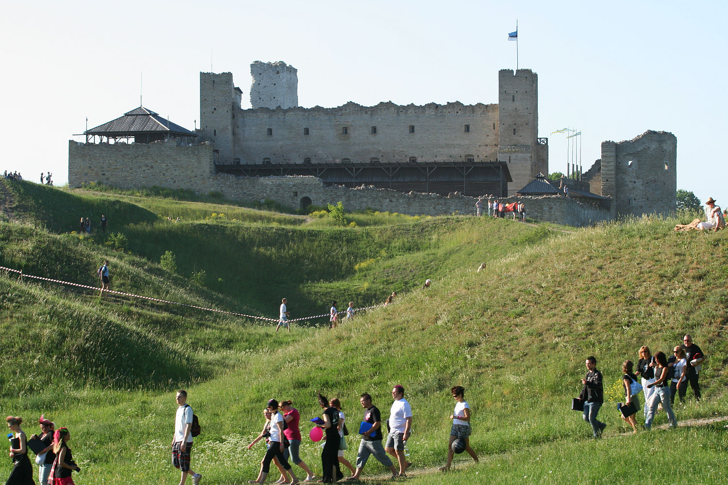 A group of people walks along a path through a grassy hillside toward a large, medieval stone castle with multiple towers, reminiscent of scenes from Laulupidu celebrations. Some people are visible on a higher path closer to the castle. One tower has a flag at the top, and the sky is clear and blue in the background.
