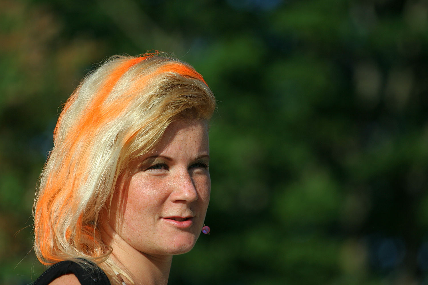 A woman with blonde hair featuring bright orange streaks stands outdoors, exuding a punk vibe. She is wearing a black top and has a small pink earbud in her left ear. The blurred background of green foliage and sunlight adds to the Laulupidu-like atmosphere. She has light skin, freckles, and an expression suggesting she is speaking or listening.