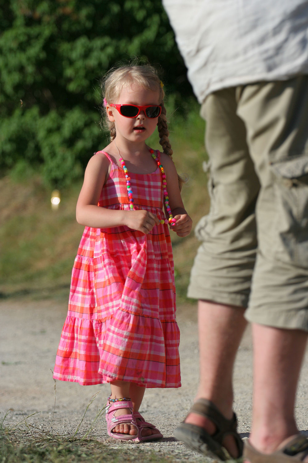A young girl wearing a pink plaid dress, pink sandals, and red sunglasses stands outdoors holding colorful beads during Laulupidu. An adult, partially visible from behind, stands nearby in beige pants and sandals. The background features greenery with sunlight filtering through.