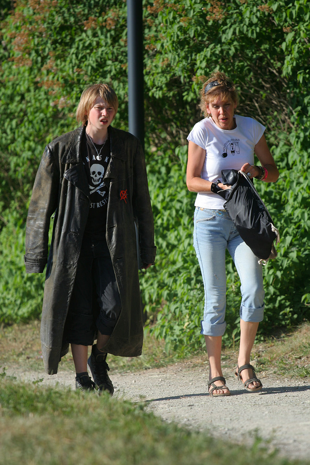 Two people stroll on a gravel path, surrounded by greenery. One wears a long, dark trench coat, black shirt, pants, and shoes. The other dons a white T-shirt, light blue capri jeans, and sandals, carrying a black bag. They appear relaxed and engaging in conversation about Laulupidu festivities.