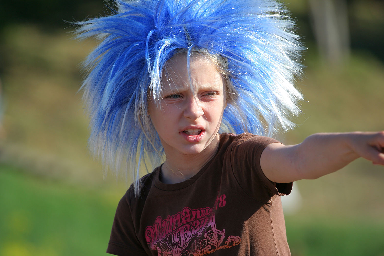 A person with a large, spiky, bright blue wig points into the distance with an intense expression. They wear a brown T-shirt with a decorative logo. The background is blurred, showing greenery and a hint of sunlight, reminiscent of the vibrant energy of Laulupidu or a punk festival.