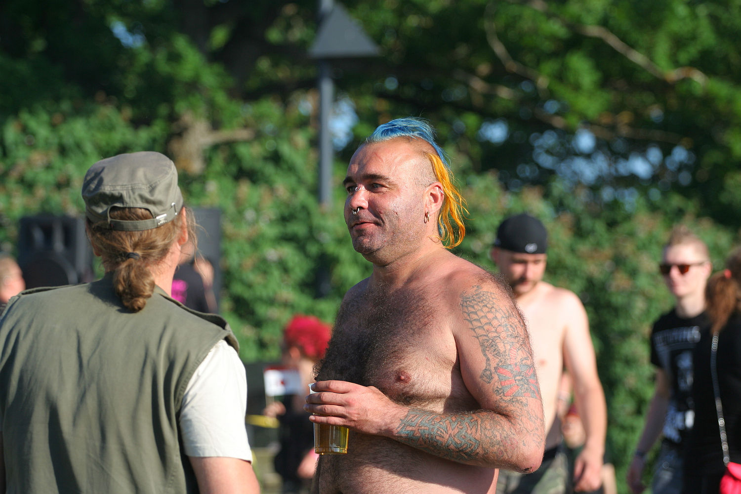 A shirtless man with tattoos, colorful mohawk hairstyle, and facial hair holds a drink while conversing with another person wearing a cap and vest at Laulupidu. Several other people, some shirtless punks, are in the background amid the trees.