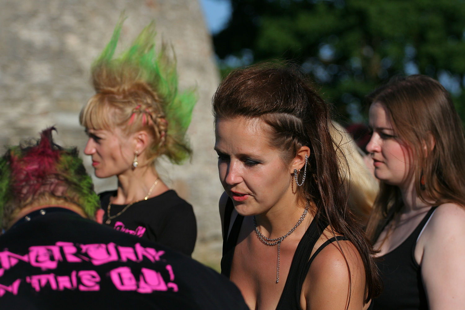 Four people are gathered outside on a sunny day. The person facing away has bright pink and green spiked hair and a black shirt. Three women face forward: one with a green mohawk, one with a brown ponytail, and one with long brown hair. Their punk style is evident from their casual outfits and visible piercings.