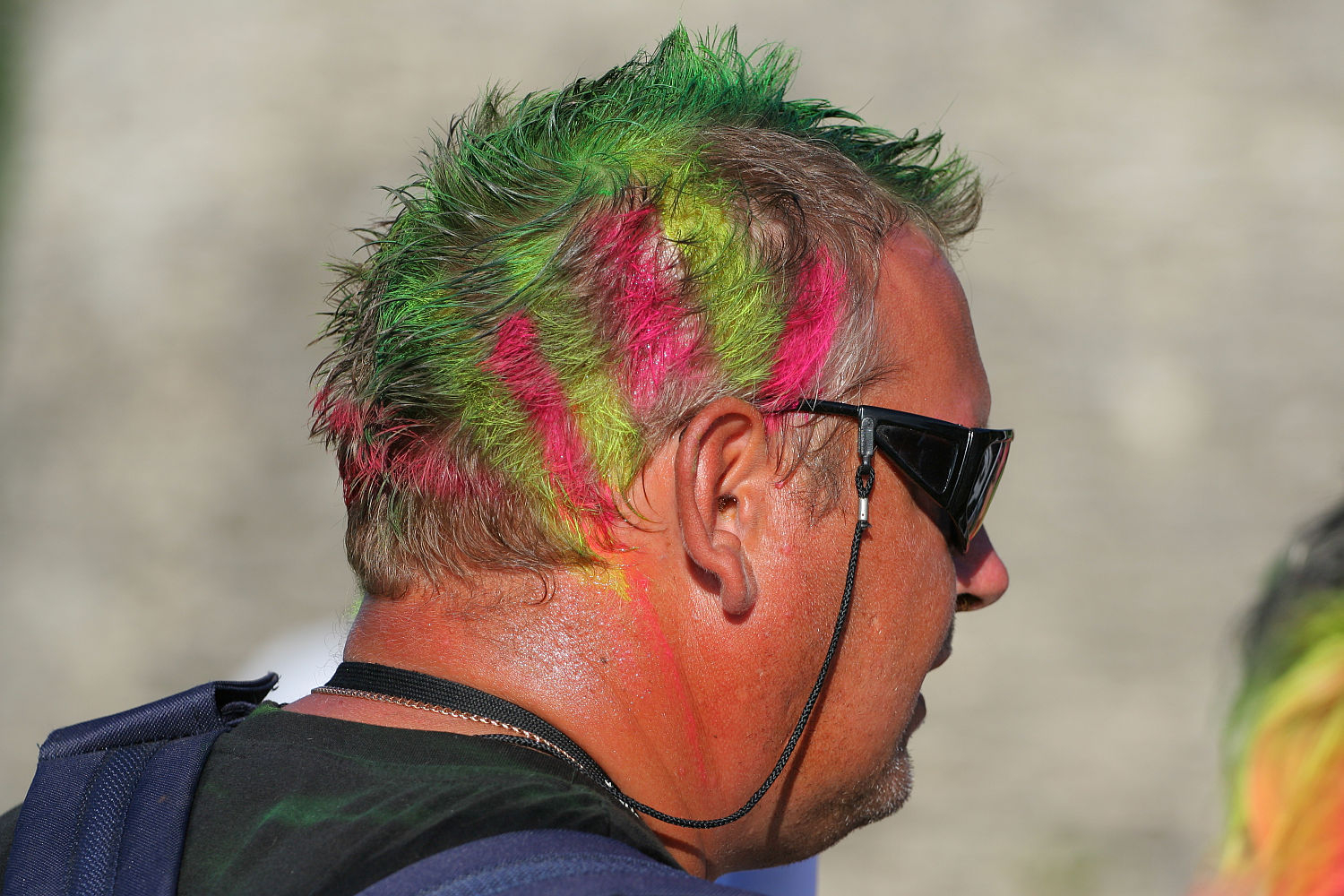 A man with short hair dyed bright green and streaked with vivid pink stands facing away. He wears dark sunglasses, and his neck and part of his face exhibit hints of orange paint, giving him a punk vibe. The background is blurred and neutral-toned, providing no additional context.