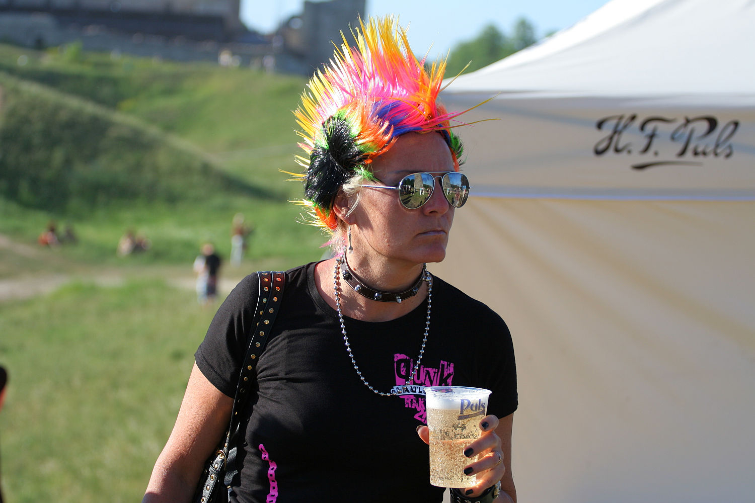A punk woman sporting a vibrant rainbow mohawk, sunglasses, and a black choker is holding a plastic cup of beer. She is wearing a black T-shirt with pink and white text. In the background, at Laulupidu, there is a white tent with writing and a grassy area with people in the distance.