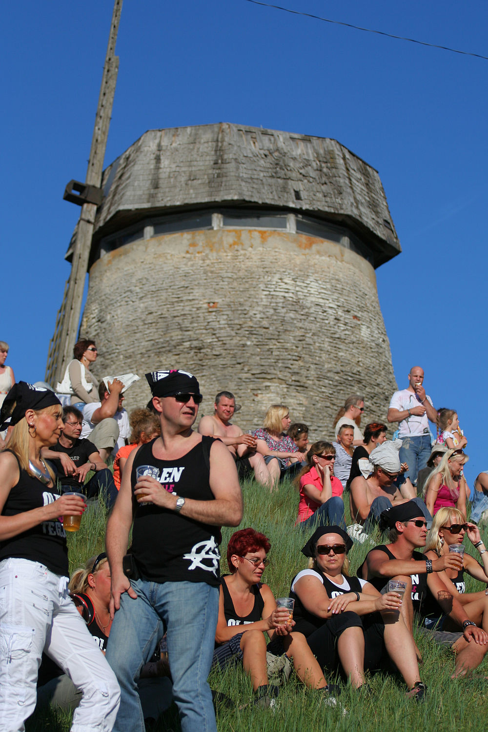 A group of people, many wearing matching black and white tank tops and baseball caps, sit and stand on a grassy hill, enjoying drinks. Behind them is a stone tower structure with a wooden roof against a clear blue sky. Some have sunglasses, others hold cups while conversing in a laid-back punk vibe at Laulupidu.
