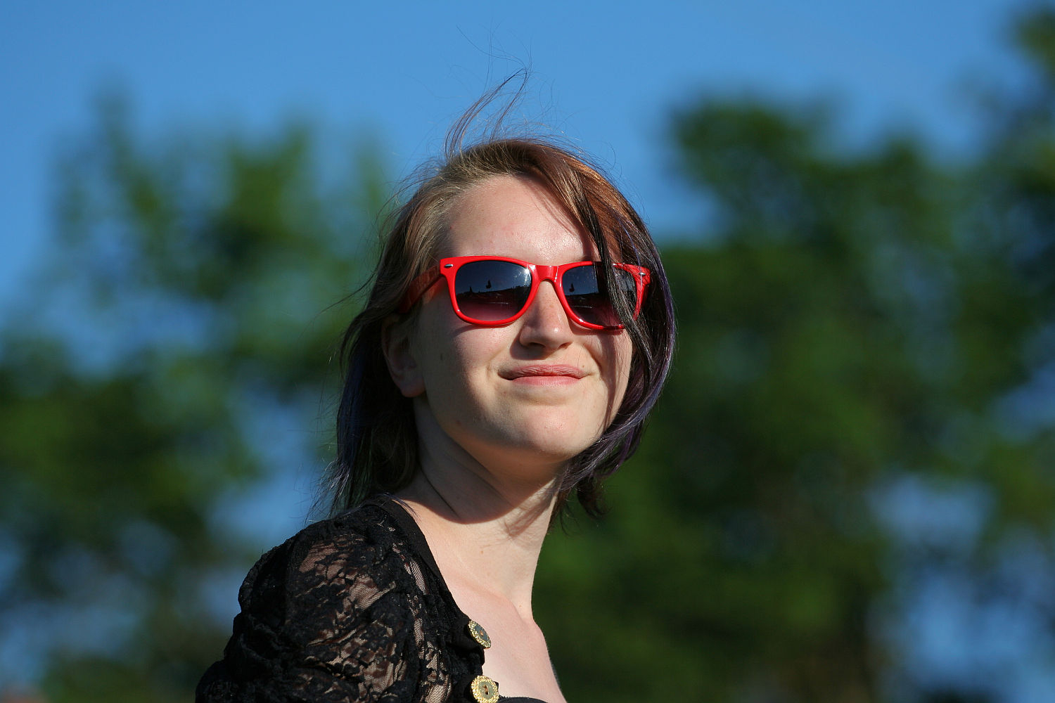 A person with shoulder-length hair wearing bright red sunglasses is smiling slightly while standing outdoors. Dressed in a black lace top with gold buttons, they exude a punk vibe. The background features blurred, green, leafy trees against a clear blue sky, reminiscent of a vibrant Laulupidu scene.