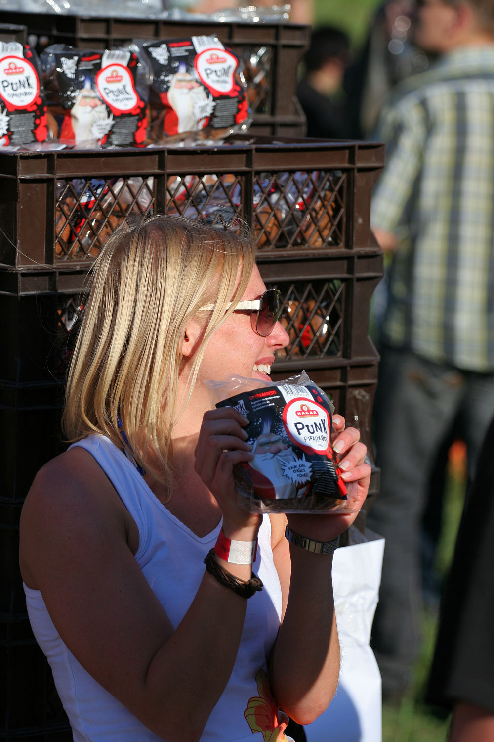 A woman with blonde hair and sunglasses, wearing a white sleeveless shirt, is holding a package of "PUNK" coffee beans and smiling. She stands next to a stack of brown plastic crates filled with additional packages of the same coffee. The background features blurred people and green grass, evoking the spirit of Laulupidu.