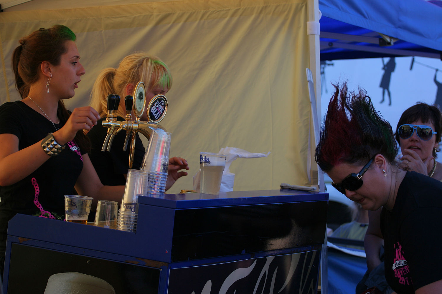 A punk woman with a green and red mohawk bends down dramatically near a bar setup, while two women in black shirts stand behind the counter, one operating beer taps. Another person watches on from the right, under a blue tent. The scene feels reminiscent of Laulupidu, set at an outdoor event or festival.