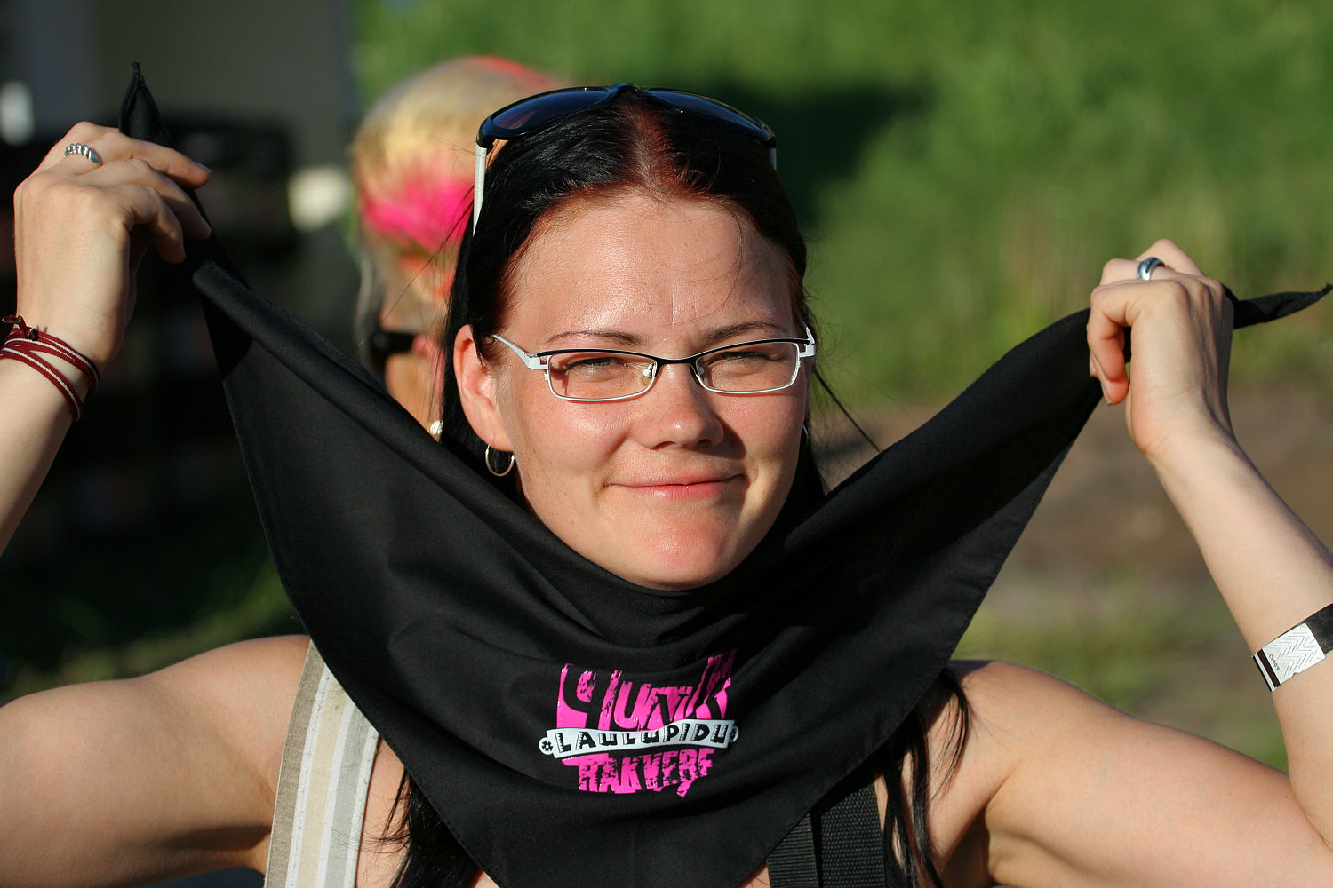 A smiling woman with glasses and dark shoulder-length hair holds a black bandana with pink writing under her chin. She is outdoors in a sunny environment, possibly at a Laulupidu festival, with greenery in the background. She wears a punk-inspired silver watch on her left wrist and has her nails painted a light color.