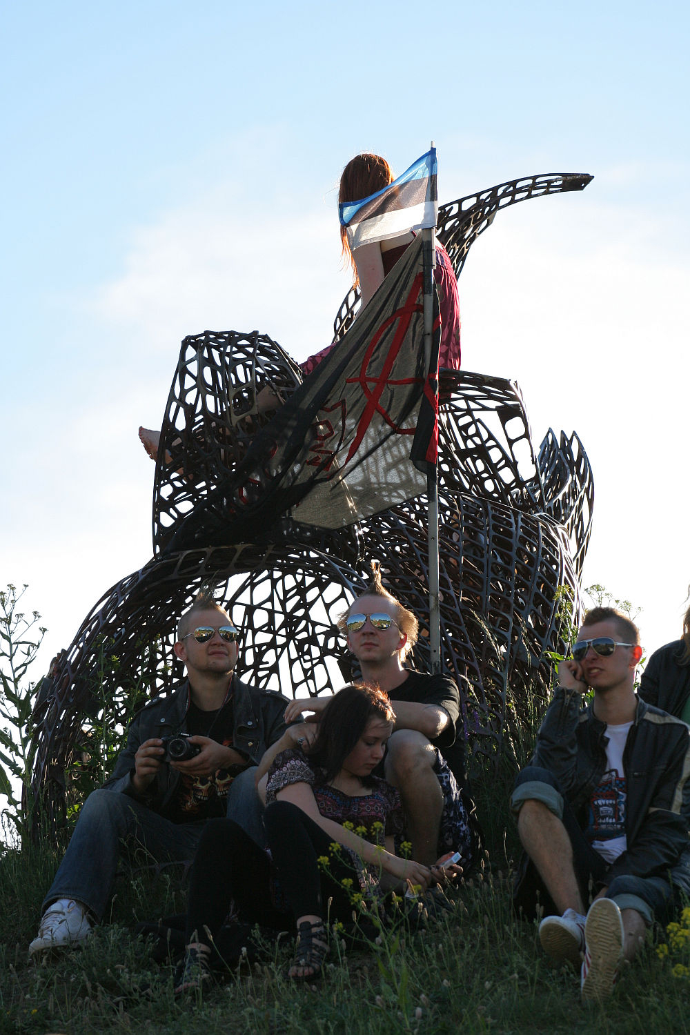 A group of young people, embracing a punk aesthetic, lounges around a large, abstract metal sculpture in a grassy area. One person is perched atop the structure, holding a flag that echoes the Estonian Laulupidu spirit with an X on a white field and bottom resembling the Estonian flag. The sky is clear and bright.