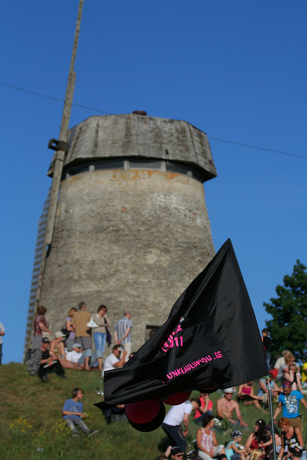 People are gathered on a grassy hill in front of an old, weathered windmill with a wooden structure at the top. A black flag with pink and white text is prominently waving in the foreground, lending a punk vibe to the scene. The sky is clear and blue, indicating a sunny day reminiscent of Laulupidu celebrations.