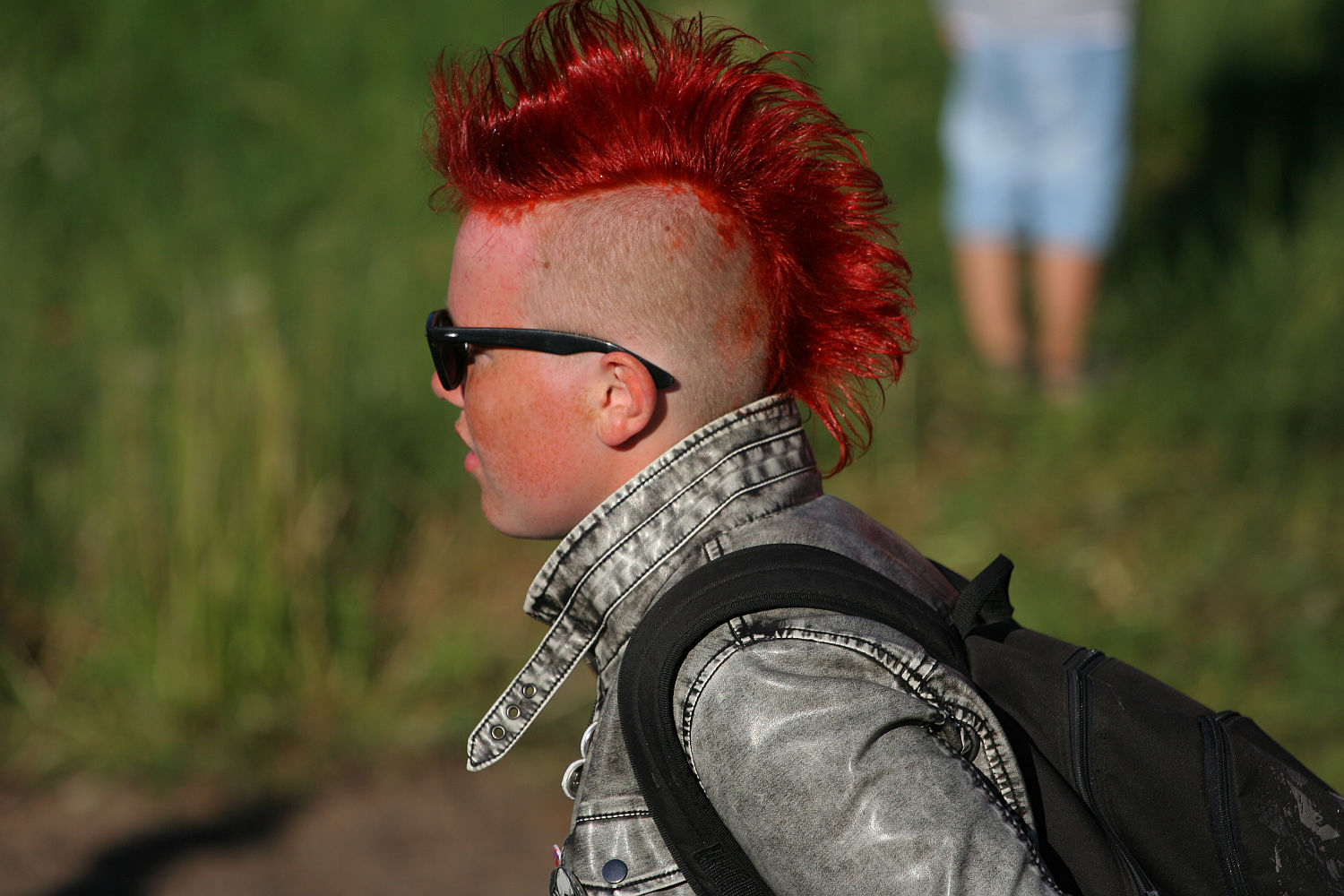 A person with a bright red mohawk hairstyle is wearing black sunglasses and a faded gray denim jacket, embodying punk style. They are photographed in profile, facing left, with a black backpack and a green, blurred outdoor background. Another person in light clothing is partly visible in the background, perhaps heading to Laulupidu.