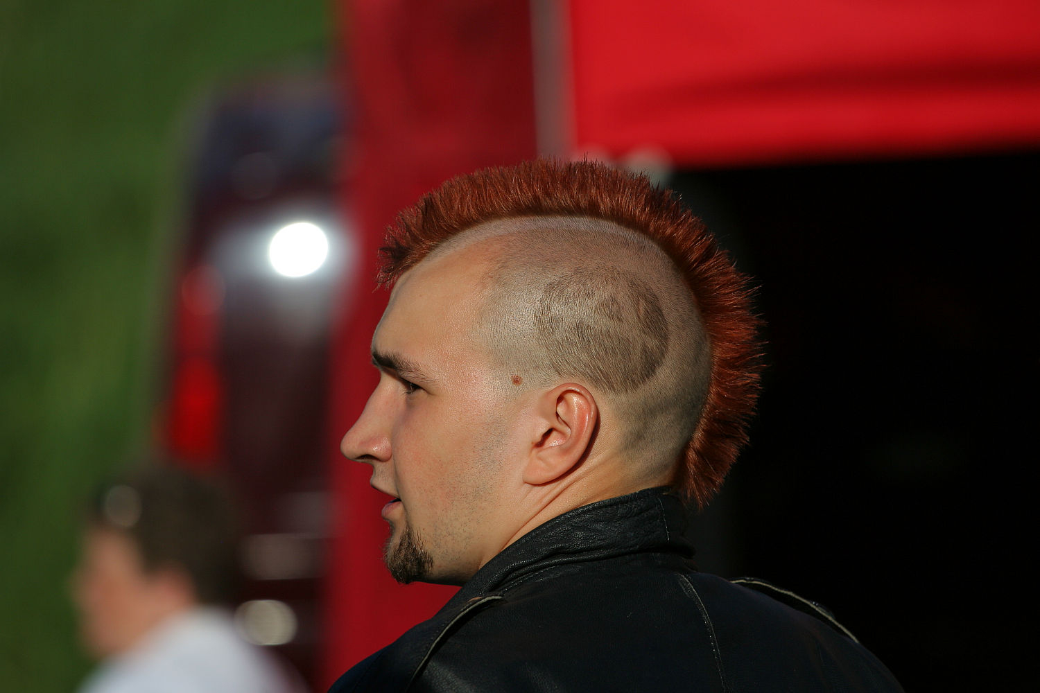 A man with a red mohawk and shaved sides stands outdoors, embracing his punk style. He has a short boxed beard and is wearing a black leather jacket. The background features a red structure reminiscent of Laulupidu festival stages and blurred greenery.