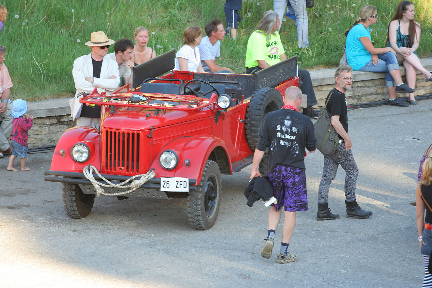 A group of people gathers around a vintage red open-top vehicle, which has a spare tire mounted on the side. Some are seated inside while others stand nearby on a concrete surface, embodying a laid-back punk vibe. The backdrop features greenery and a stone wall, with more individuals sitting on the grass in the background, reminiscent of Laulupidu's communal spirit.