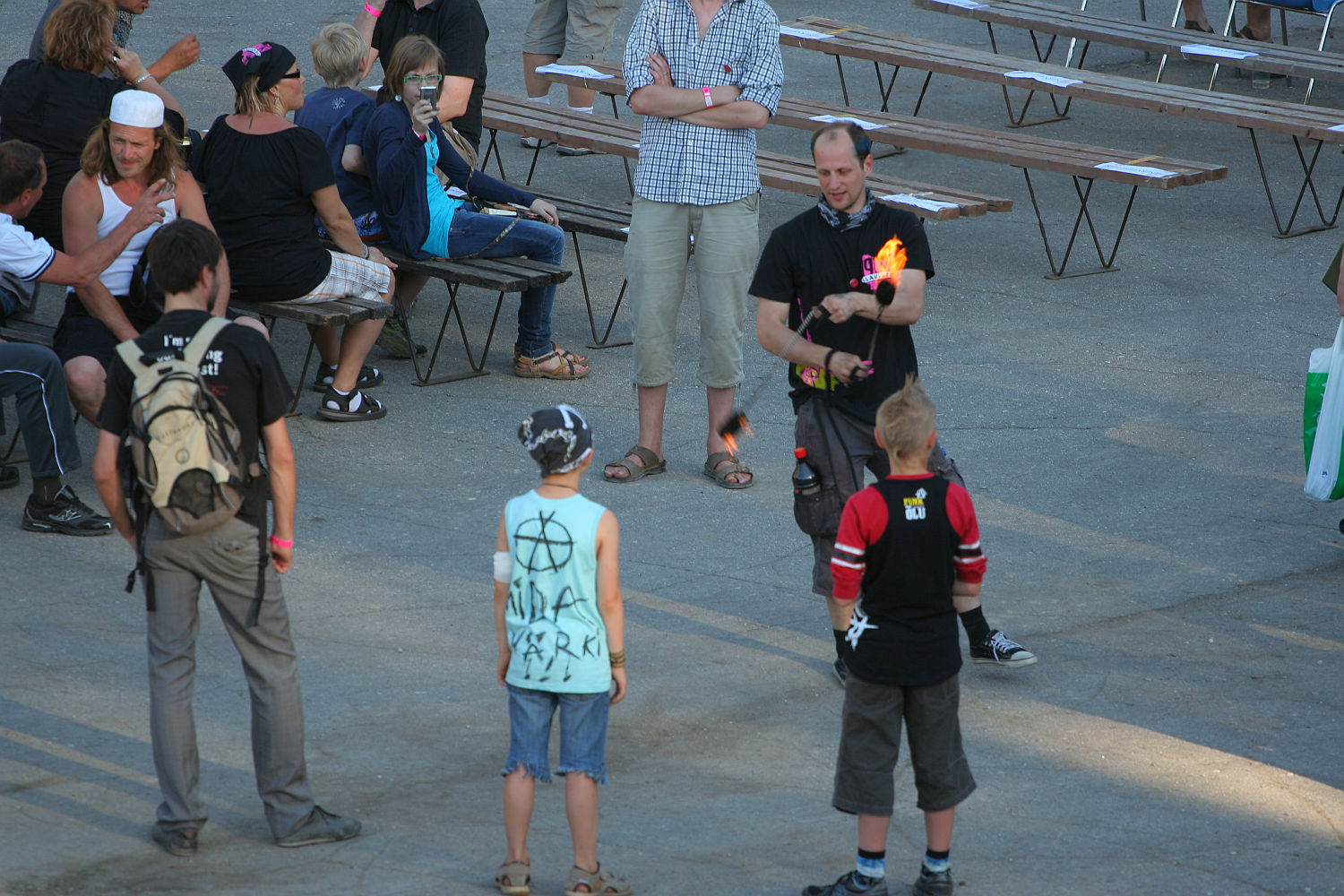 A person dressed in black is juggling flaming torches while three young people with a punk style watch, standing in a circle around the performer. In the background, other people are sitting or standing near wooden picnic tables, engaging in conversation or observing the performance.
