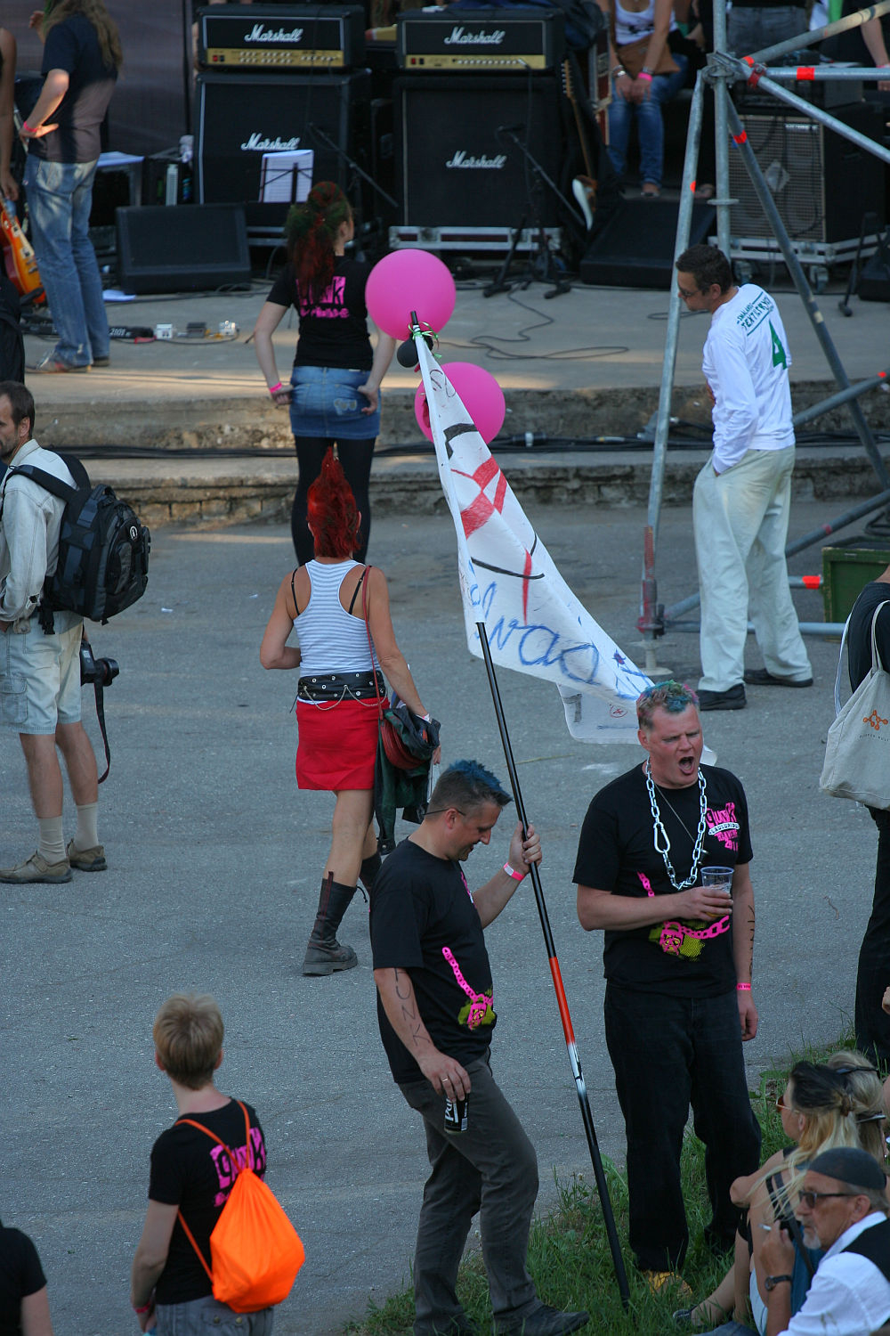 A group of people at an outdoor event blends punk and tradition. One person with a red mohawk and red skirt walks away holding pink balloons. Another person in front holds a large white flag with writing. Several individuals wear black shirts with bright pink text. Stage equipment and speakers are visible in the background, reminiscent of Laulupidu's vibrant atmosphere.