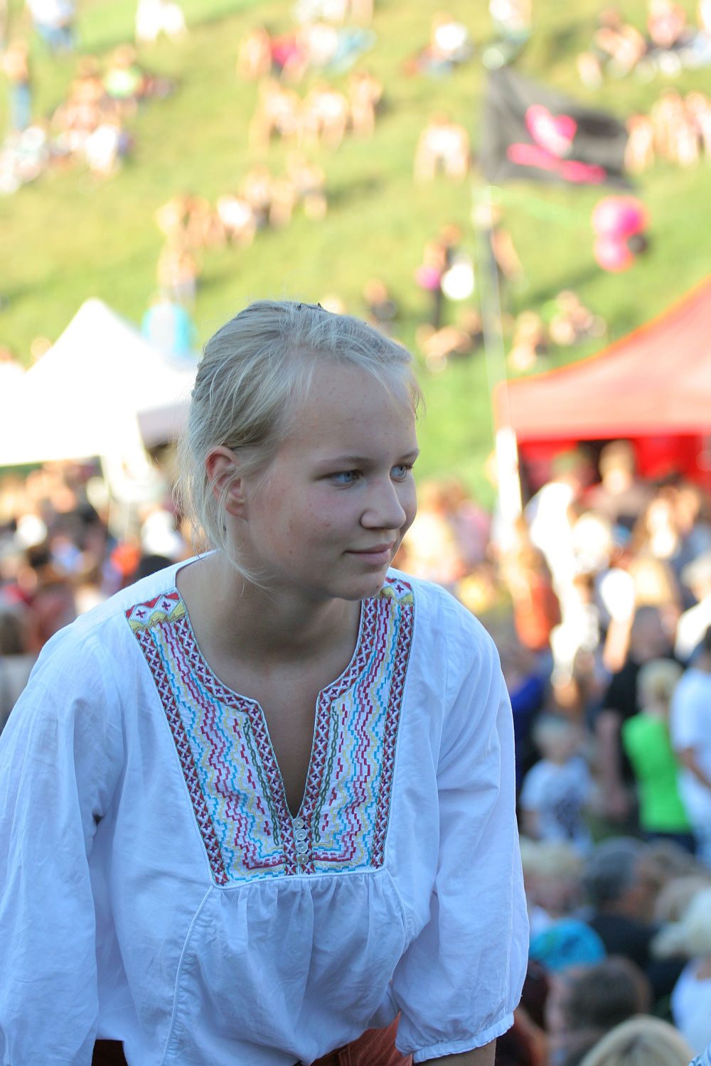 A young woman with blonde hair tied back stands outdoors at a crowded Laulupidu event. She is wearing a white blouse with colorful, intricate embroidery around the collar. People and tents with vibrant colors are blurred in the background, indicating a lively atmosphere. The setting appears to be a festival or fair on a grassy hill.