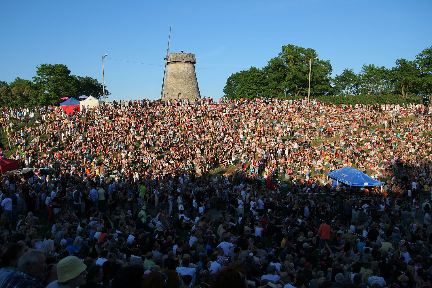 A vast crowd of people gathered on a grassy, sloping hillside under a clear blue sky, with a windmill in the background. The crowd is densely packed, all facing the stage area at the bottom of the slope where Laulupidu performances infuse traditional melodies with a hint of punk flair. Various tents and canopies, including a blue one, are dotted around.