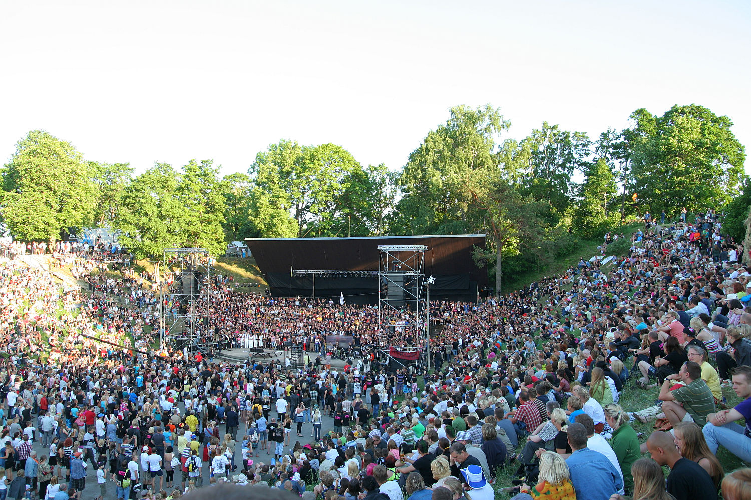 A large outdoor amphitheater, reminiscent of Laulupidu, is filled with a crowd of spectators on a bright, sunny day. The audience surrounds a central stage where an energetic punk performance is taking place. Green trees and a clear sky are visible in the background, adding to the festive atmosphere.