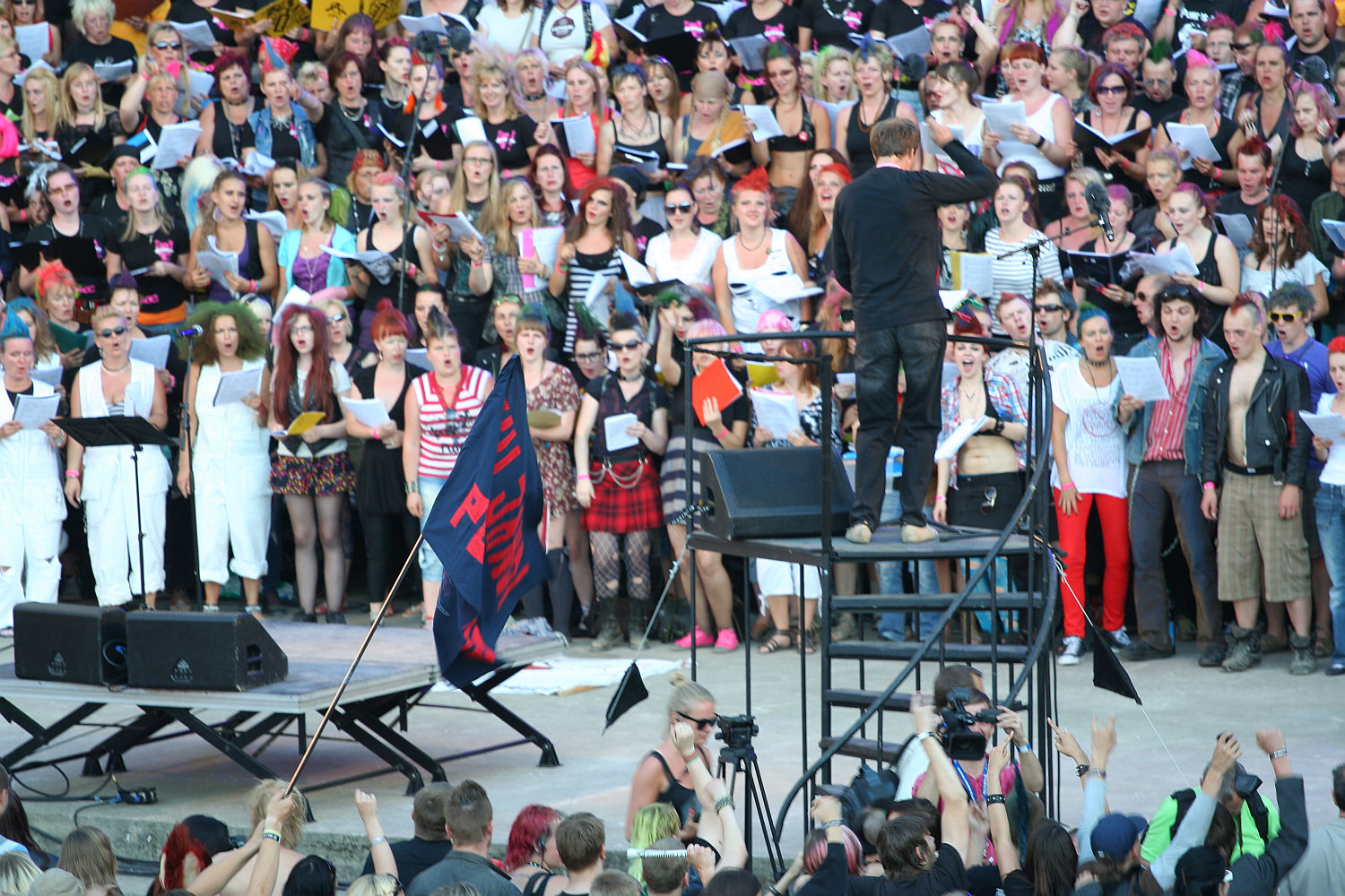 A large choral group, many in colorful costumes and accessories, performs outdoors. A conductor stands on a black platform, leading the group. Below him on the left, a sign reads "I wanna rock!" There's an audience in the foreground and various equipment and crew members visible. It's like a punk-themed Laulupidu!