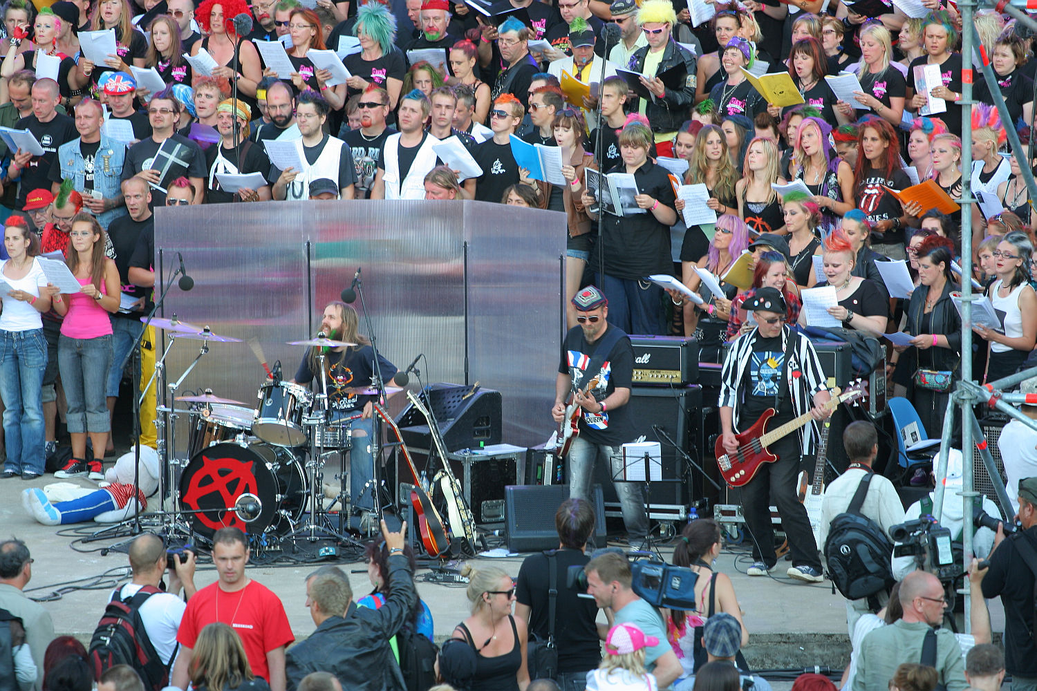 A punk rock band performs in front of a large, diverse crowd at an outdoor event reminiscent of Laulupidu. The band members play drums, guitar, and bass while people in the audience enthusiastically watch and sing along. The backdrop is filled with individuals holding song sheets, creating a lively, engaging atmosphere.