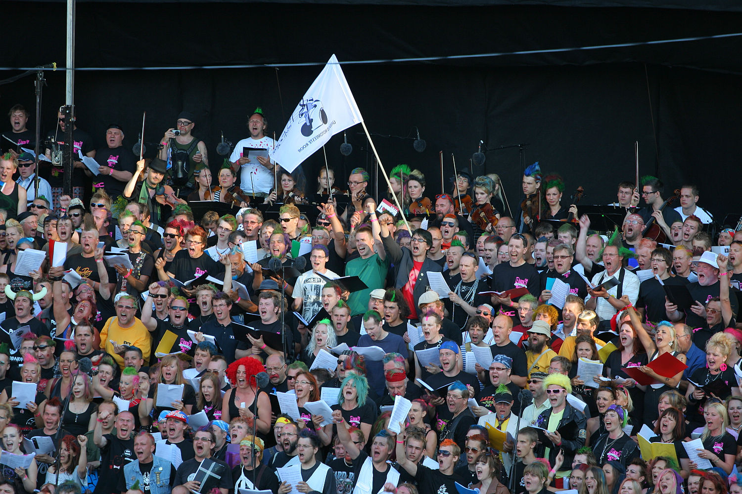 A large, diverse group of people sing in a choir, with some holding colorful folders and wearing vibrant costumes reminiscent of a Laulupidu festival. One person in the center holds a white flag with a black illustration. The dark background contrasts their enthusiasm and engagement, adding a touch of punk flair.