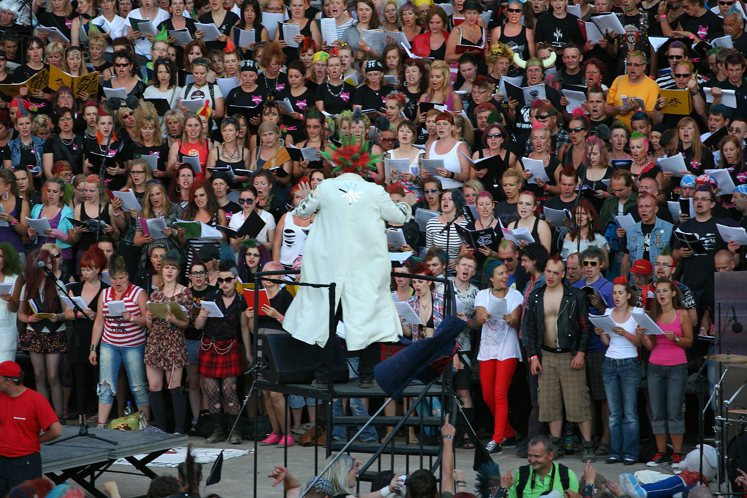 A conductor in a white coat and red spiked wig leads a large choir of diverse individuals at Laulupidu. The choir members are holding music sheets and appear engaged in singing. The scene is vibrant with a variety of punk outfits and hairstyles among the singers. The event takes place outdoors.