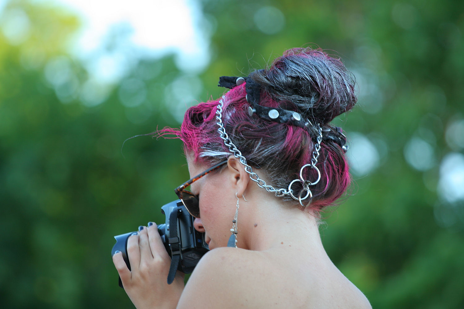 A person with dark pink and black hair styled in a punk updo decorated with chains and pins is holding a camera up to their face. They are wearing sunglasses, earrings, and a strapless top. The background is out of focus green foliage.