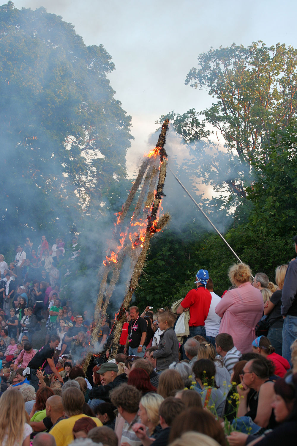 A large crowd, reminiscent of a Laulupidu festival, gathers on a grassy hill, watching as a tall wooden structure with geometric patterns is set ablaze. Smoke rises from the flames. People of various ages are present amidst the trees with green foliage, some holding flowers and displaying subtle punk vibes.