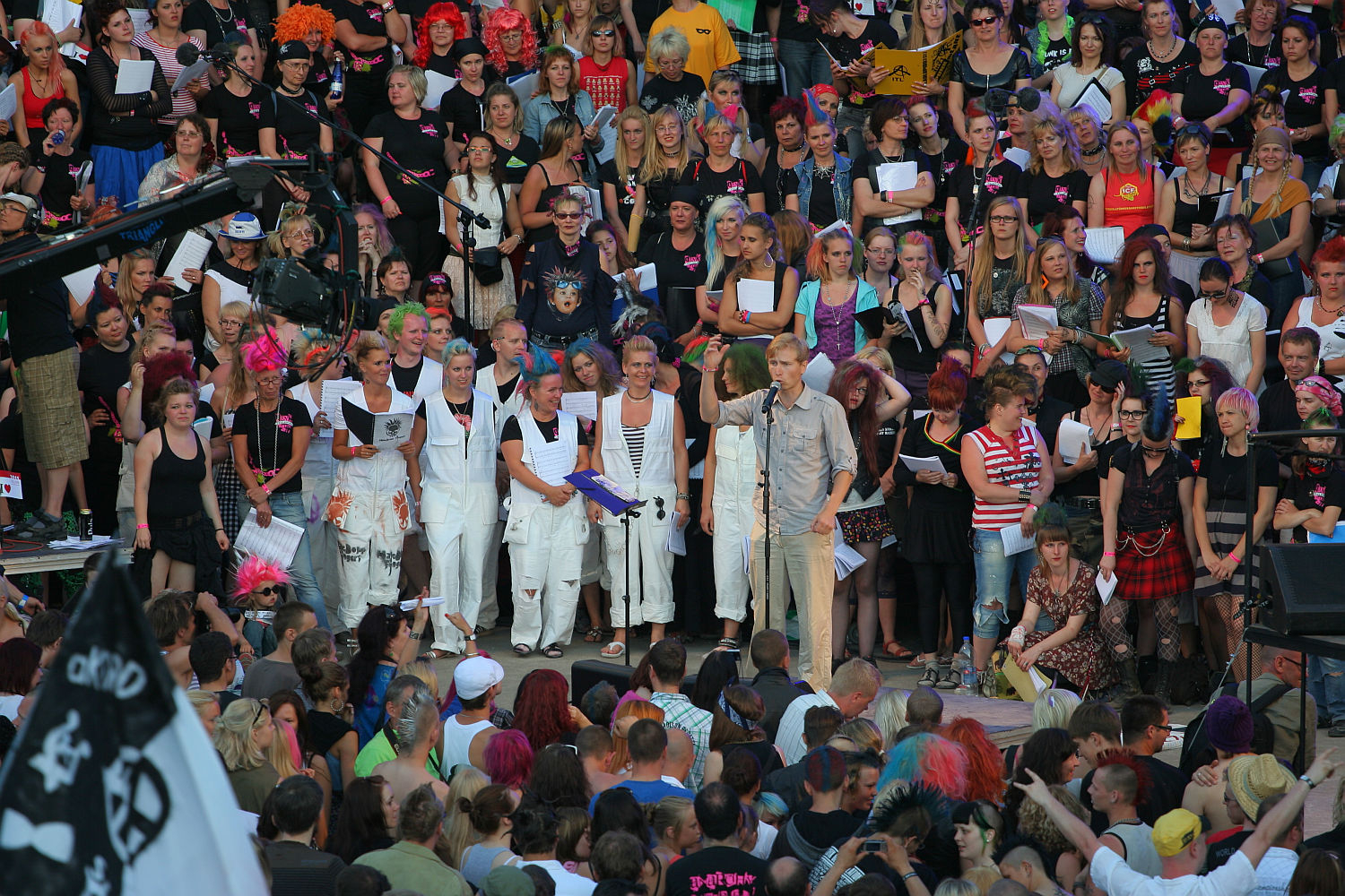 A large, colorful crowd is gathered outdoors, some dressed in bright costumes and wigs. The atmosphere echoes a spirited Laulupidu festival. At the center, a person in a gray shirt stands out against a group of people dressed in white. The lively scene has people holding papers, cheering, and some taking photos.
