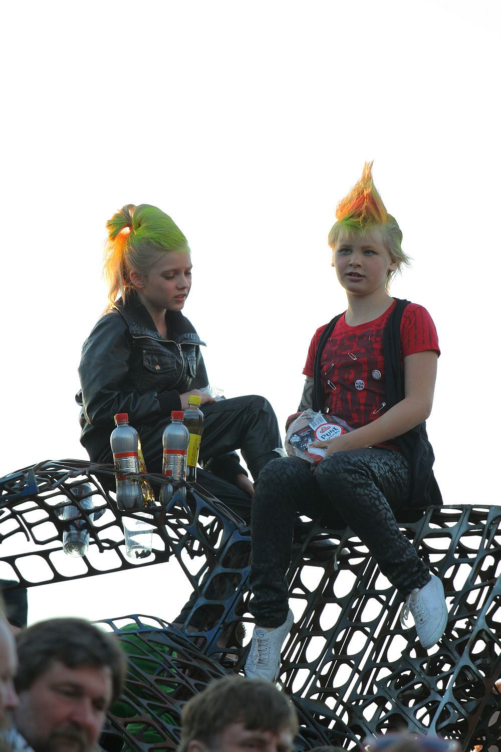Two children with bright green and orange mohawk hairstyles sit atop a metal structure. Each child holds a bottle with beverage, and both have serious expressions. The child on the left wears a black leather jacket, while the one on the right sports a red and black outfit, capturing the essence of punk at Laulupidu.