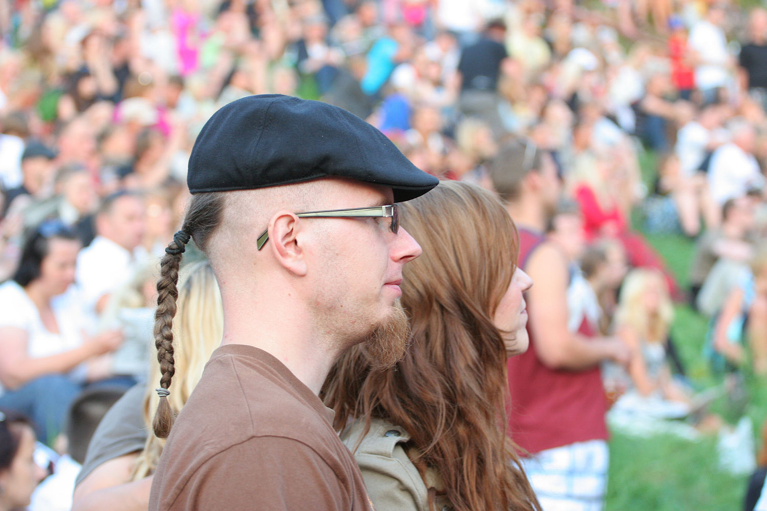 A man with a braided ponytail, glasses, and a black cap stands next to a woman with long brown hair in a crowd at an outdoor Laulupidu event. The background shows many people sitting on grass, creating a lively and social atmosphere with hints of punk fashion scattered throughout.
