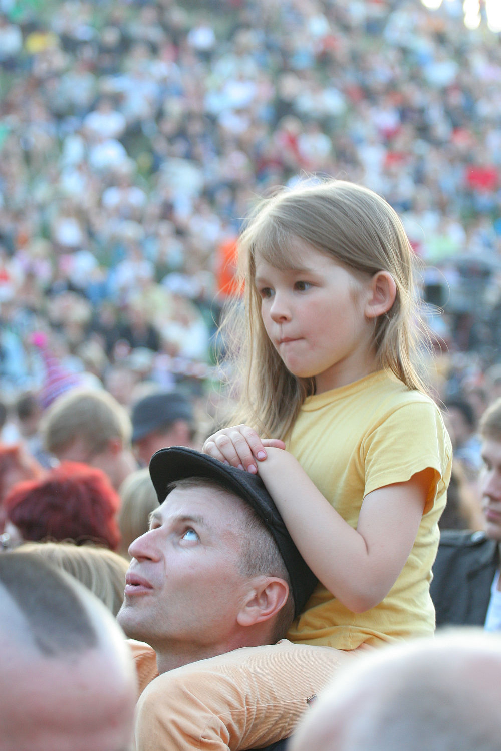 A man wearing a hat carries a young girl on his shoulders at Laulupidu, a vibrant outdoor event. The girl, dressed in a yellow shirt, looks ahead with a thoughtful expression. In the background, the large crowd has a slightly punk vibe, enhancing the energy of the scene while remaining blurred to emphasize the foreground.