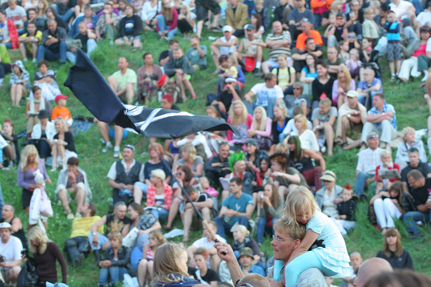 A large crowd is seated on a grassy hillside at an outdoor event. In the foreground, a man with a young child on his shoulders waves a black flag featuring a white "X" and a skull and crossbones symbol. It feels like Laulupidu meets Punk, with people dressed in casual summer clothing, including t-shirts, shorts, and hats.