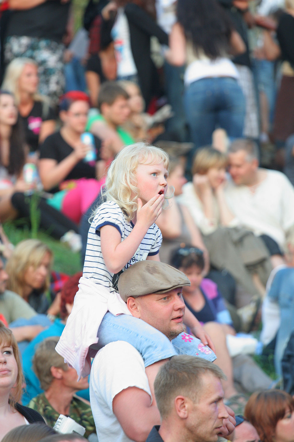 A young girl with blonde hair, wearing a striped shirt, sits on a man's shoulders in a crowded outdoor gathering reminiscent of Laulupidu. The man, donning a flat cap and white shirt with subtle punk flair, is surrounded by people sitting on grass, many engaged and holding snacks. The atmosphere is lively and social.