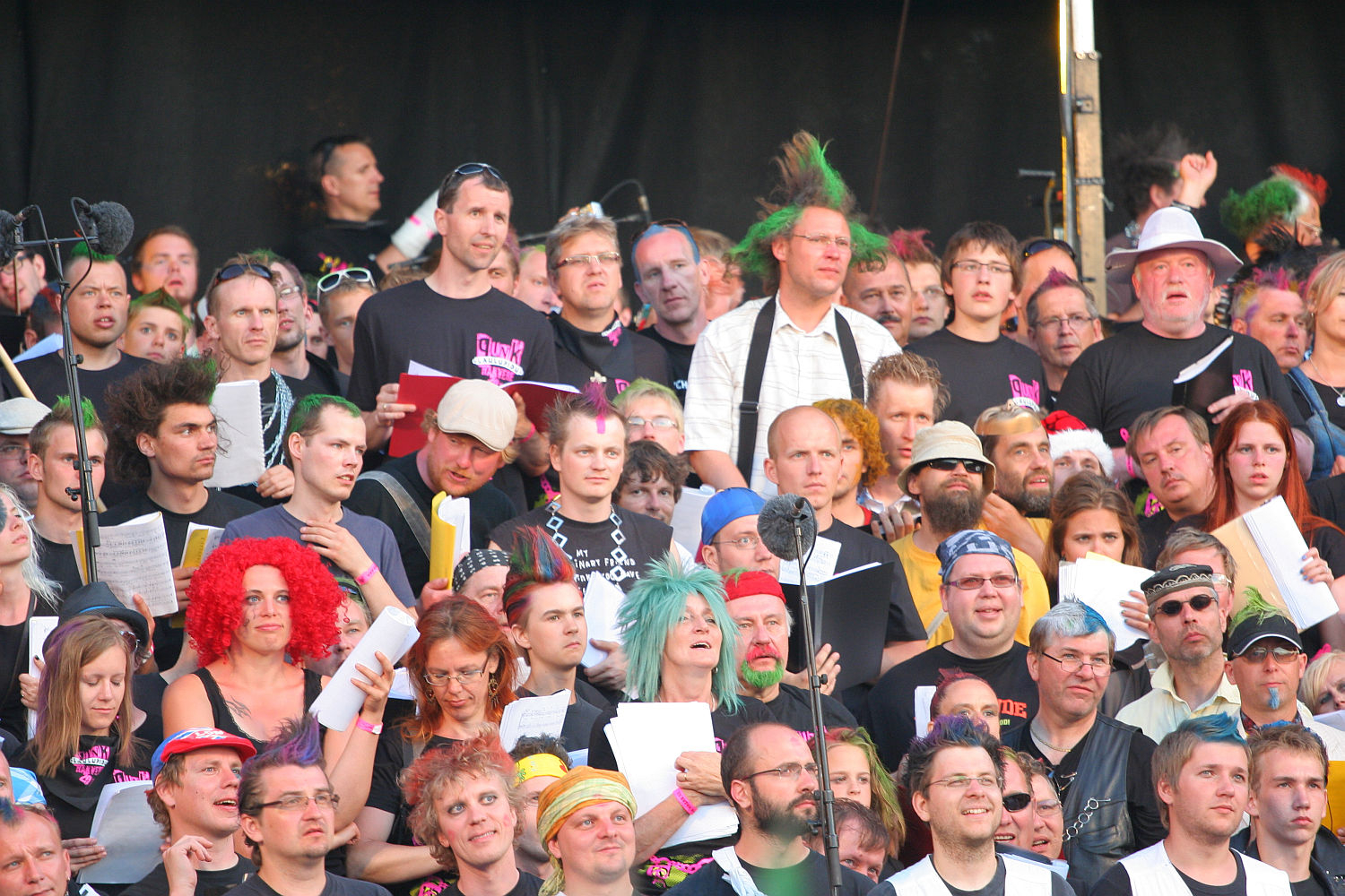A large and diverse group of people are gathered closely together, many holding sheet music and singing. They are dressed in casual clothes, with some donning colorful wigs and punk accessories. Enthusiastic and focused, they stand against a dark backdrop, possibly indicating a stage setting reminiscent of Laulupidu.