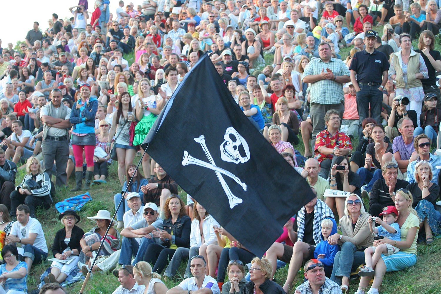 A large crowd sits on a grassy slope during Laulupidu, with people dressed in casual summer clothing. In the center, a black flag with a white skull and crossbones adds a punk flair. The audience appears engaged and diverse, featuring a mix of adults, children, and seniors.