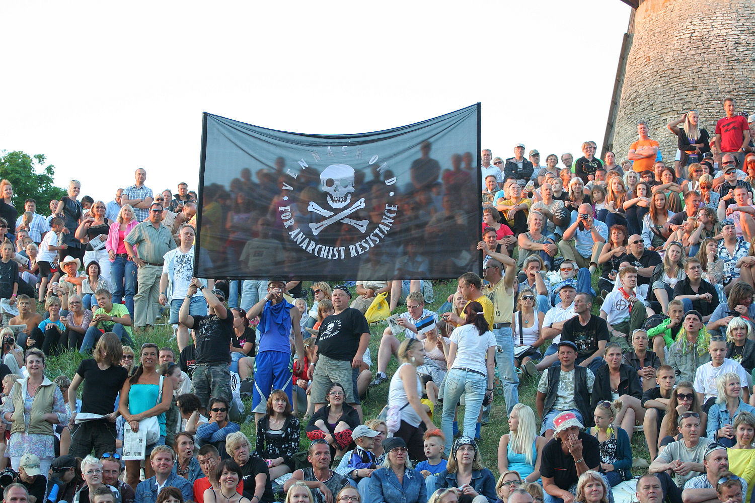 A diverse crowd is seated on a grassy slope at an outdoor gathering, reminiscent of Laulupidu. They are holding a large black flag with a white skull and crossbones design and the text "FOR ANARCHIST RESISTANCE." The punk-spirited crowd includes adults and children, all appearing engaged and attentive.