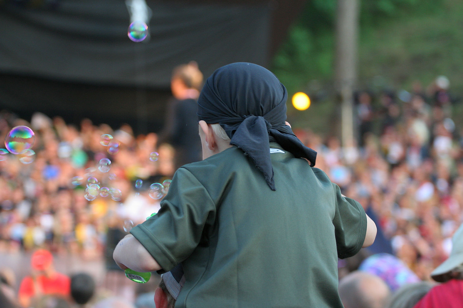 A person with a black headscarf and green shirt is seen from behind, sitting on someone's shoulders at an outdoor Laulupidu event. In the foreground, soap bubbles float around, and in the background, a large crowd of people can be observed amid a blurred punk performer on stage.