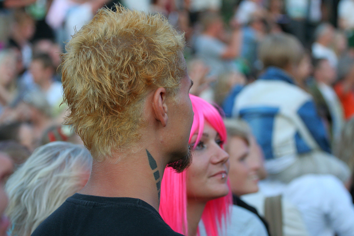 A man with bleach blonde hair and black geometric neck tattoos stands in a crowd at Punk Laulupidu, seen from behind. Beside him, a woman with a bright pink wig smiles while looking up. The background is blurred but filled with people wearing casual clothing.