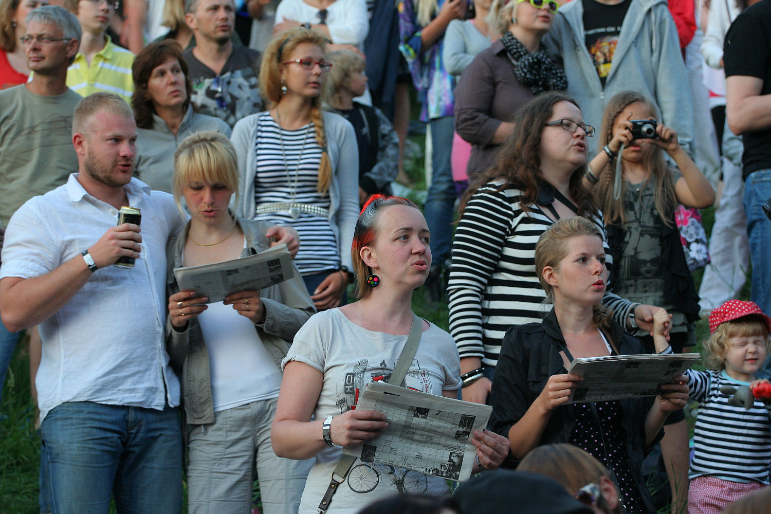 A diverse group of people are gathered outdoors, many holding newspapers. Some are standing, while others appear to be singing or reading, reminiscent of a Laulupidu celebration. The crowd includes men, women, and children in casual clothing. A few people in the background are taking photos and observing the scene.