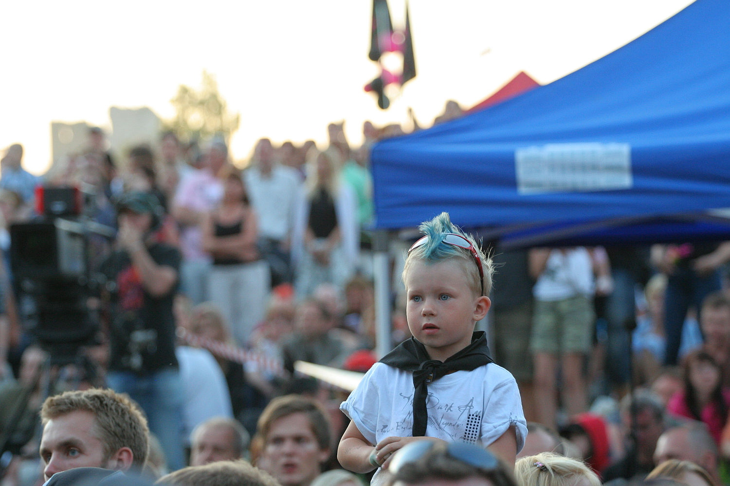 A young child with blue hair styled in a mohawk sits on someone's shoulders at an outdoor event. The child is wearing a white shirt and a black scarf. Surrounding the child is a large crowd of people under a blue canopy, suggesting a lively and energetic atmosphere reminiscent of Laulupidu.
