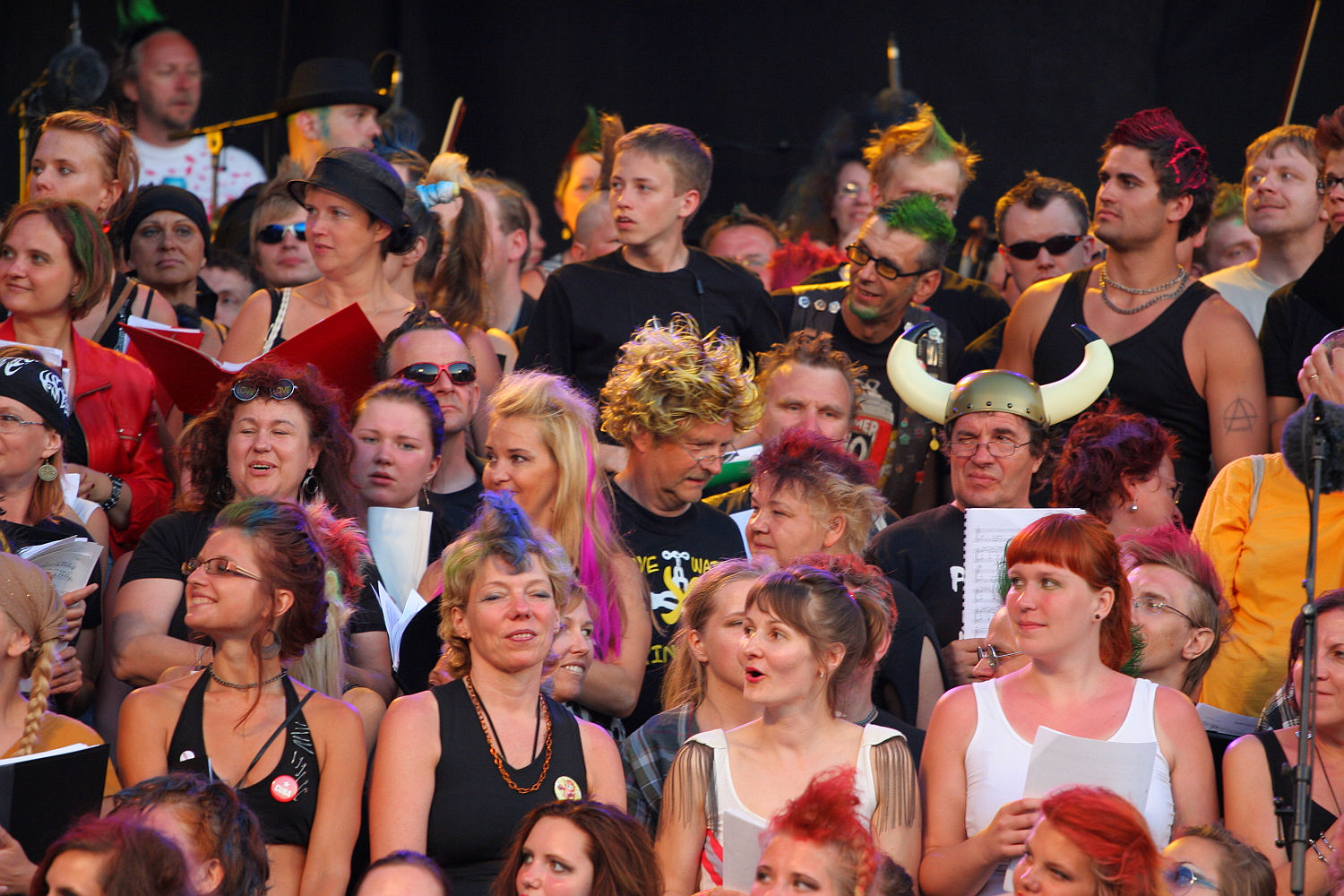 A diverse and lively crowd of people are gathered, some dressed in colorful and eclectic costumes, including Viking helmets, wigs, and face paint that add a touch of punk flair. Various expressions are visible, ranging from joyful to focused. Many hold sheets of paper, suggesting they are part of a Laulupidu chorus or performing group.
