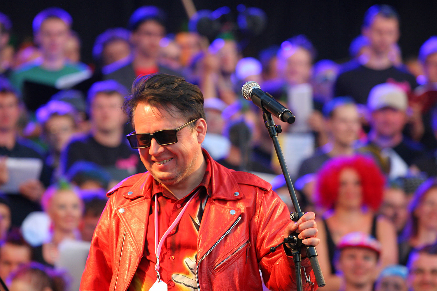 A man wearing a red leather jacket and sunglasses stands in front of a microphone on a stage. Smiling, he grips the microphone stand as if ready to deliver an electrifying punk performance. The crowd, slightly blurred, resembles a vibrant Laulupidu festival with colorful clothing, accessories, and signs.