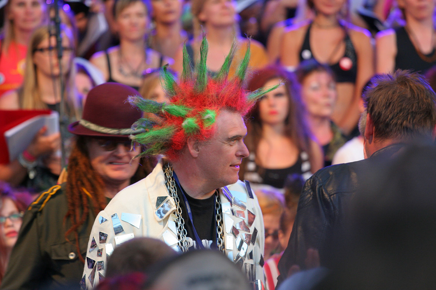 A person with spiked green and red hair, wearing a jacket with silver triangular patches and a large chain necklace, stands amidst the crowd at Punk Laulupidu. Another person with long dreadlocks and a maroon hat is near them. The background features a large group of people, some watching intently.